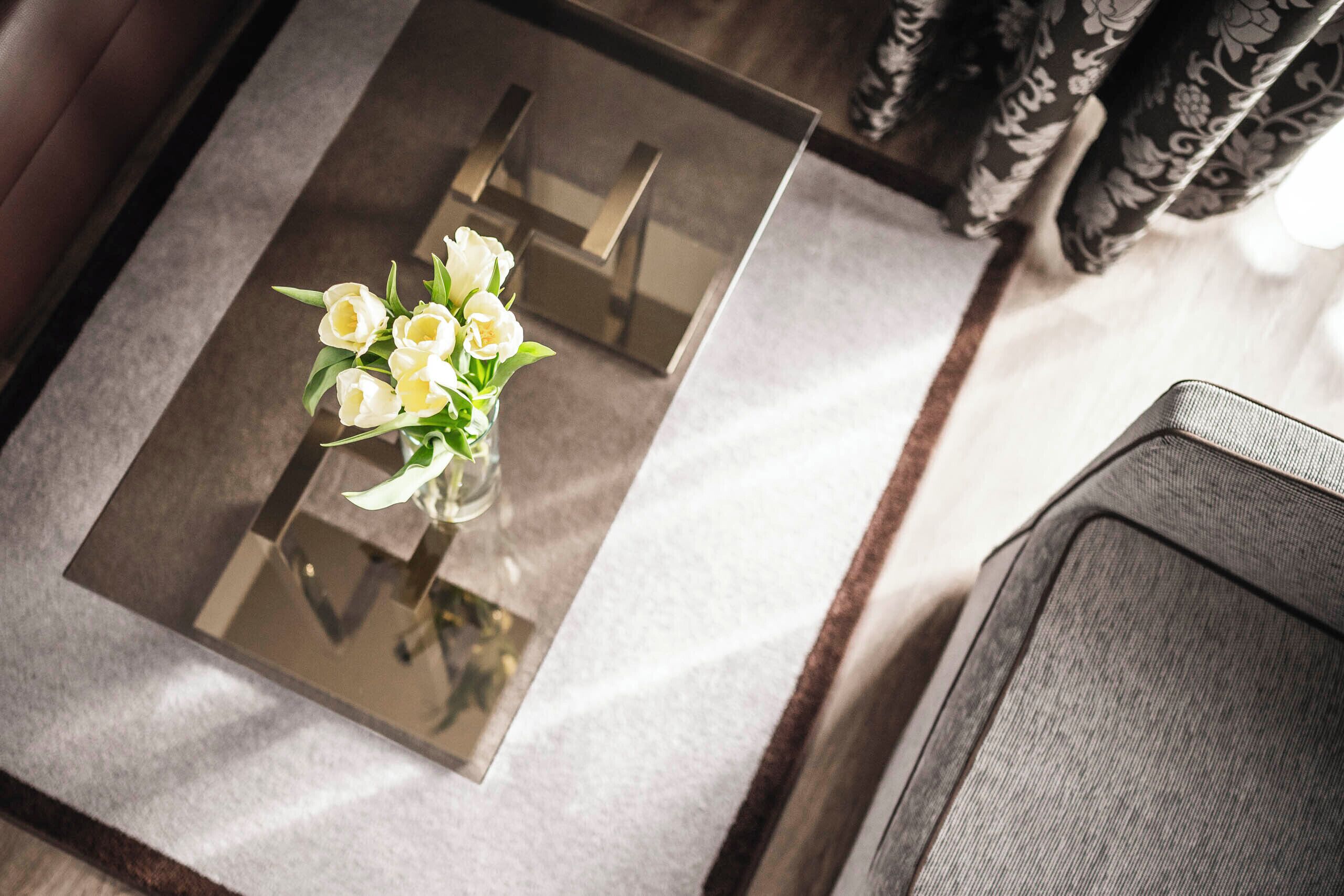 A glass-top table with a bouquet of white flowers, a chair, and patterned curtains in the background.