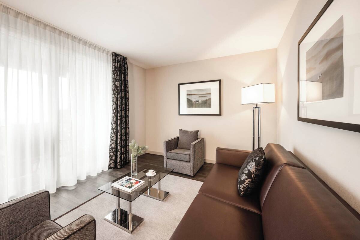 A modern hotel room with a brown leather sofa, a glass coffee table, a grey armchair, and a floor lamp.