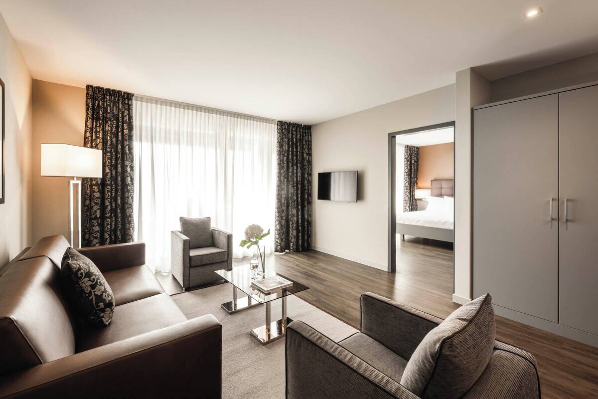 A modern hotel room with a sofa, armchair, and a glass coffee table. There is a TV mounted on the wall and a wardrobe.