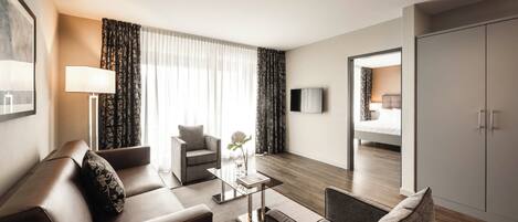 A modern hotel room with a sofa, armchair, and a glass coffee table. There is a TV mounted on the wall and a wardrobe.