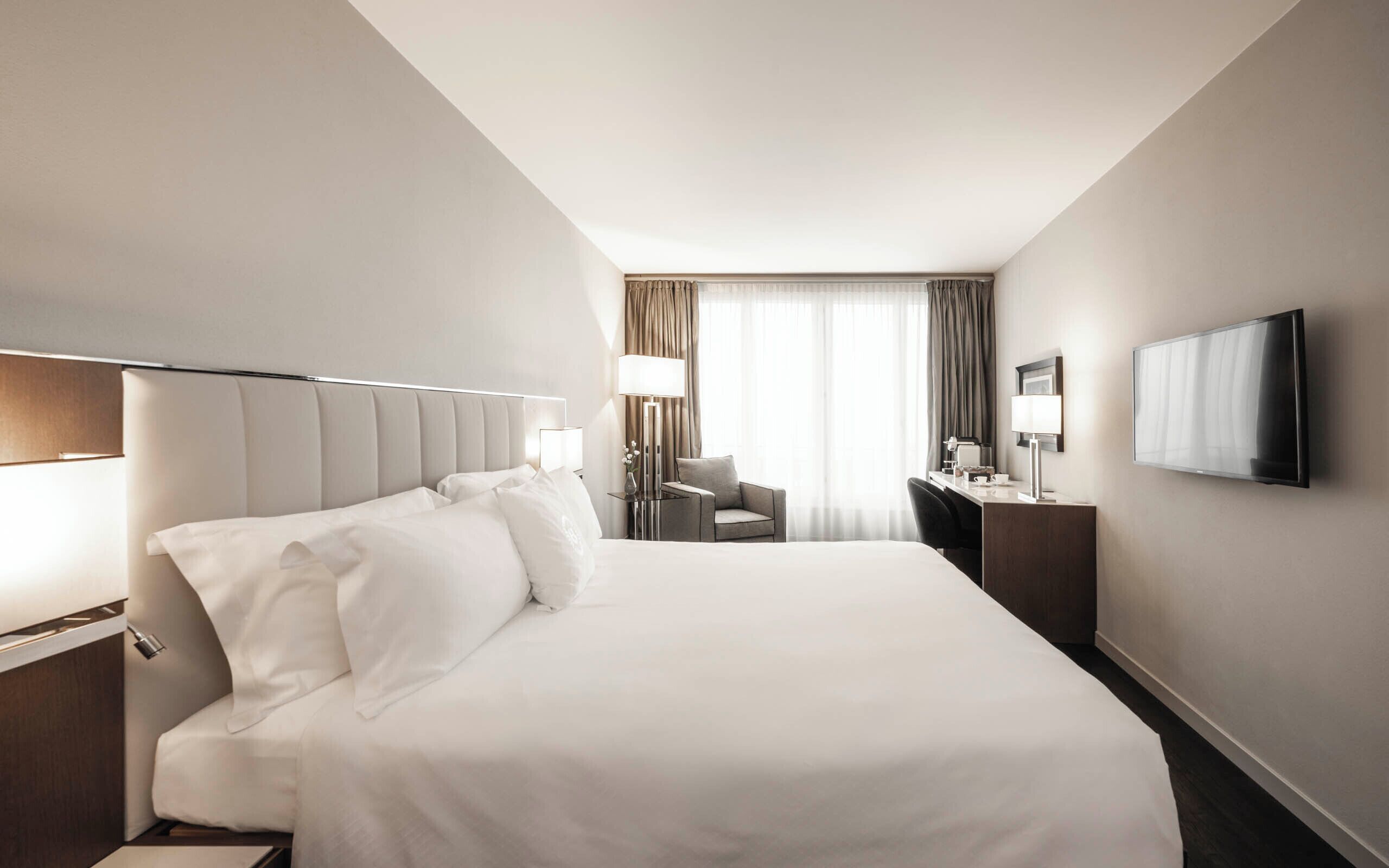 Habitación de hotel con una cama grande, un escritorio con silla, televisión y ventana con cortinas.