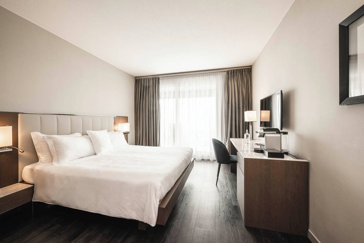 A modern hotel room with a large bed, a desk, a chair, and a TV.