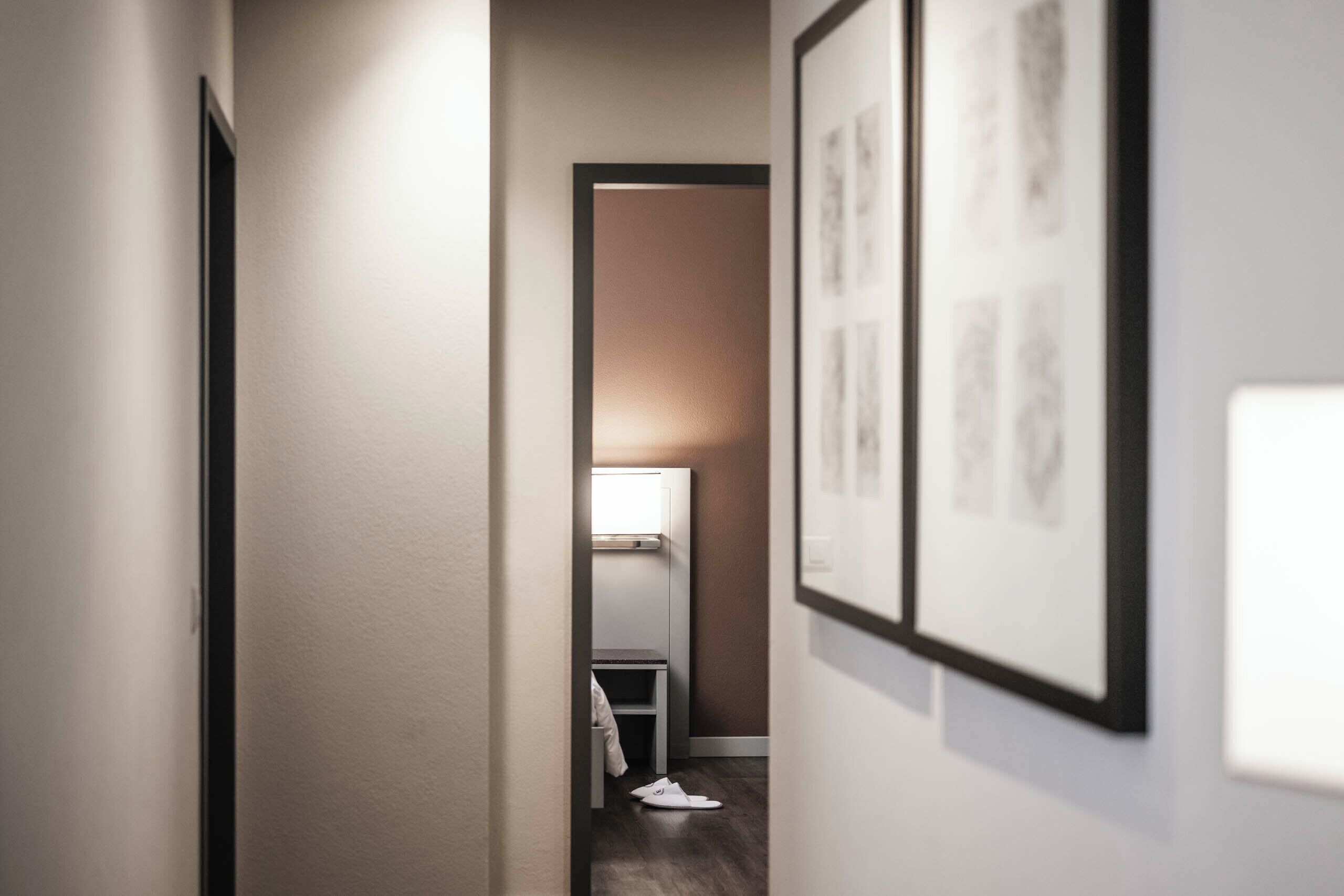 A narrow hallway with a door on the left, a framed picture on the right, and a desk with a lamp in the background.