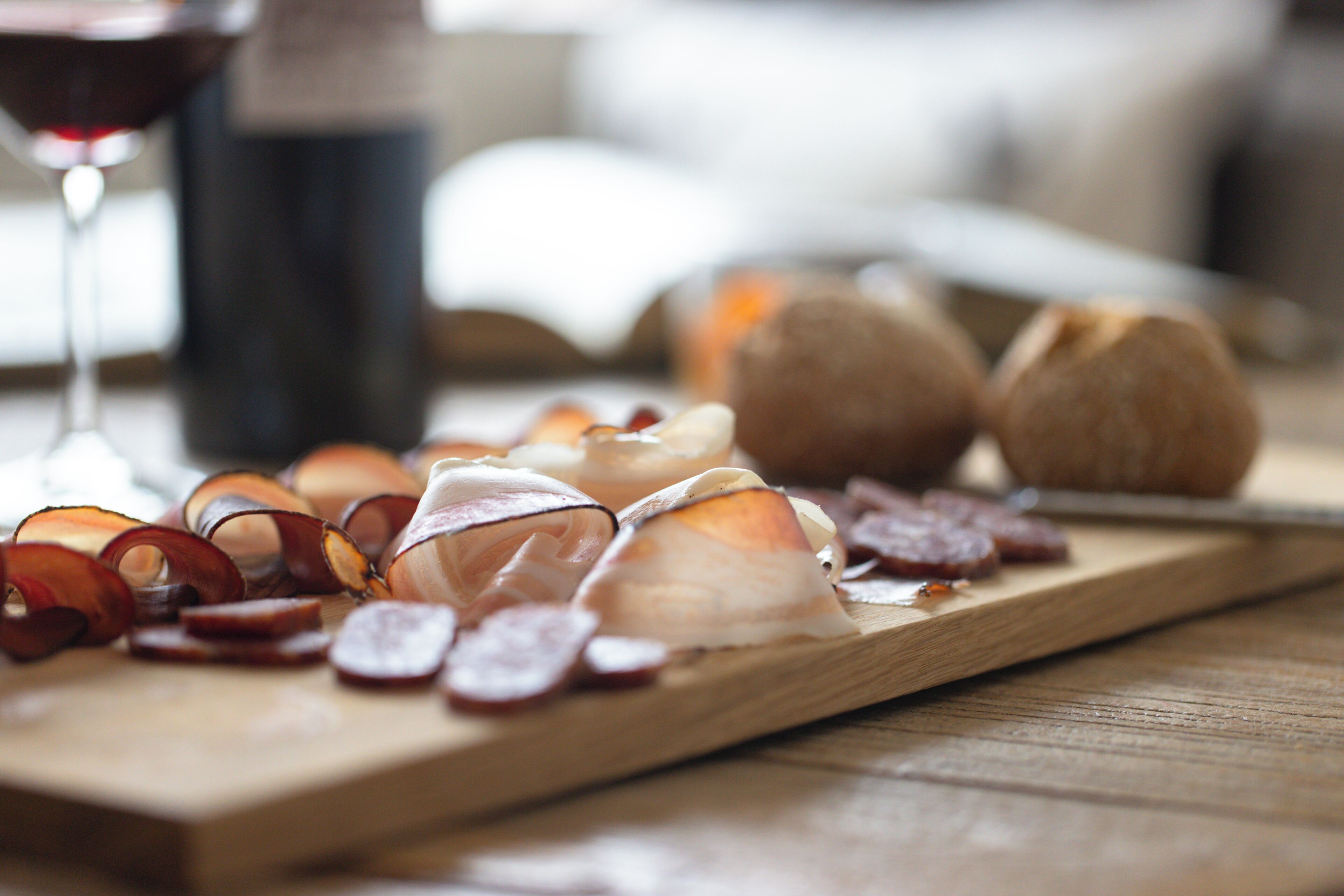 Ein Holzbrett mit geschnittenen Fleischwaren und Käse, ein Glas Rotwein und ein Messer auf einer Holztischplatte.