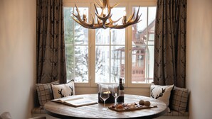A round wooden table with a book, wine glasses, and a bottle, surrounded by chairs, with a deer-patterned curtain and a chandelier.
