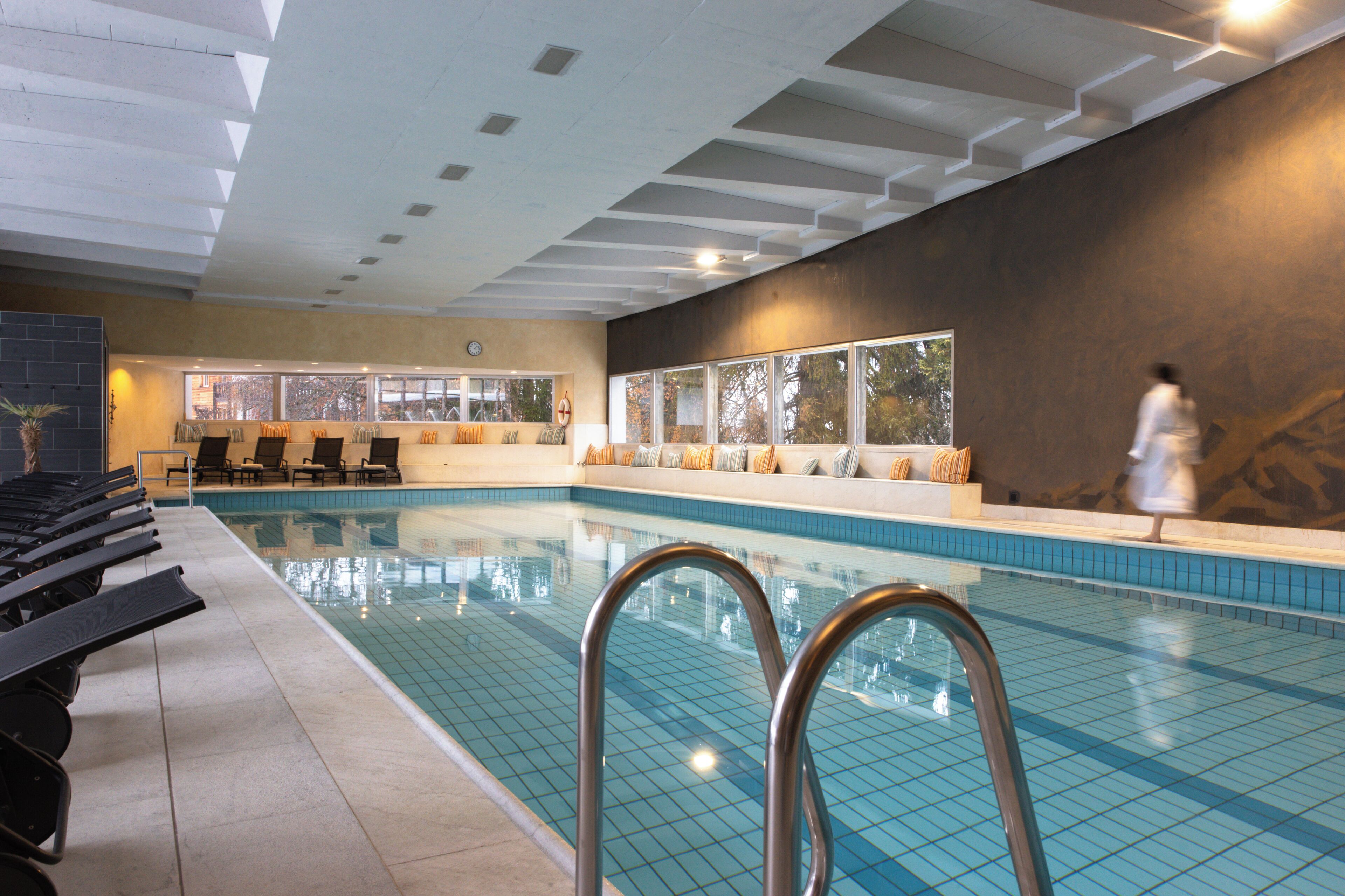 An indoor swimming pool with clear water, lounge chairs, and a view of the outdoors through large windows.