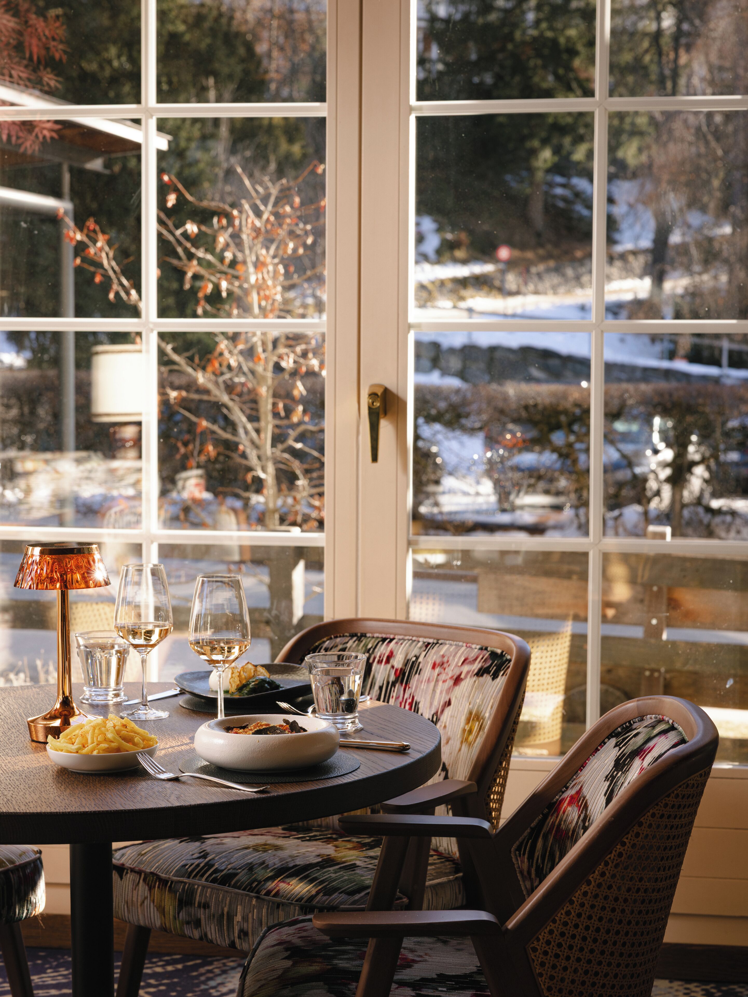 Ein Essbereich mit einem Tisch für zwei Personen, einer Lampe und Blick auf eine verschneite Landschaft durch große Fenster.
