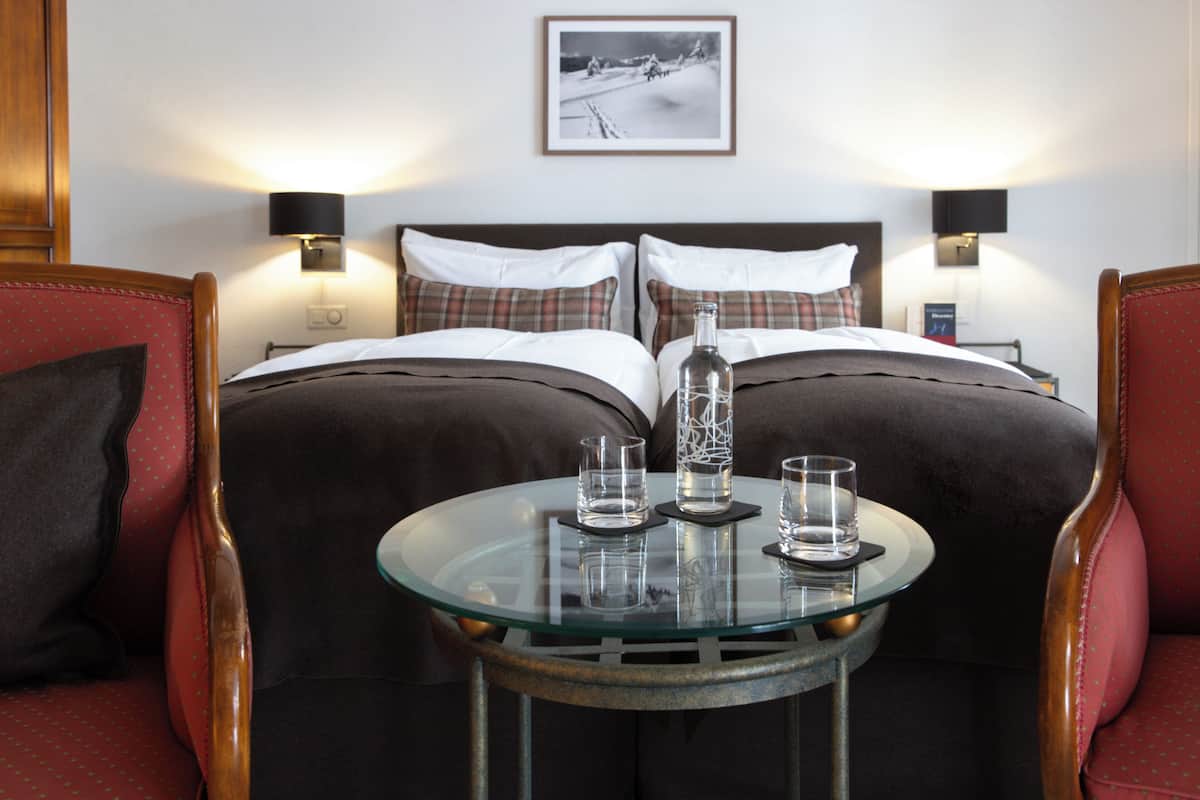 A hotel room with a bed, two bedside lamps, a glass table with two glasses and a bottle, and a framed picture on the wall.