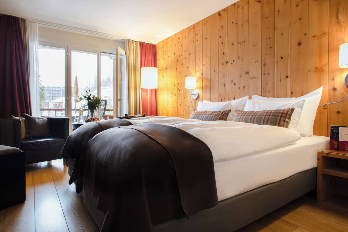 A hotel room with a large bed, wooden panel walls, a sofa, and a view through a sliding door.
