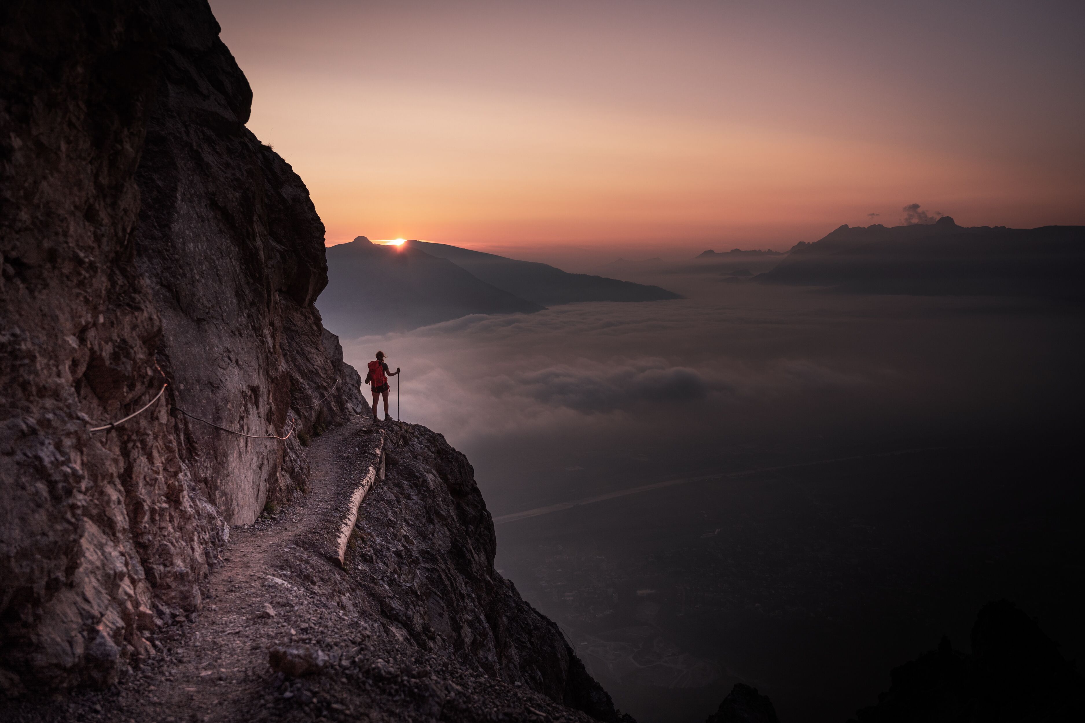 A person standing on a mountain ridge with a rope for safety, overlooking a valley with a sunrise or sunset.