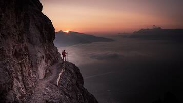 A person standing on a mountain ridge with a rope for safety, overlooking a valley with a sunrise or sunset.