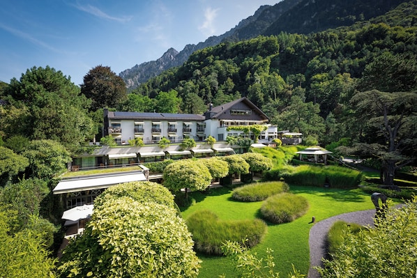 A hotel with multiple guest rooms, a garden with manicured lawns and hedges, and a mountainous backdrop.
