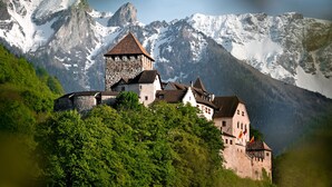 Un château de pierre historique, composé de plusieurs bâtiments, entouré d’une végétation luxuriante et avec, en arrière-plan, des montagnes aux sommets enneigés.