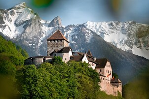 Un castillo de piedra histórico con varias construcciones, rodeado de vegetación exuberante y montañas nevadas al fondo.