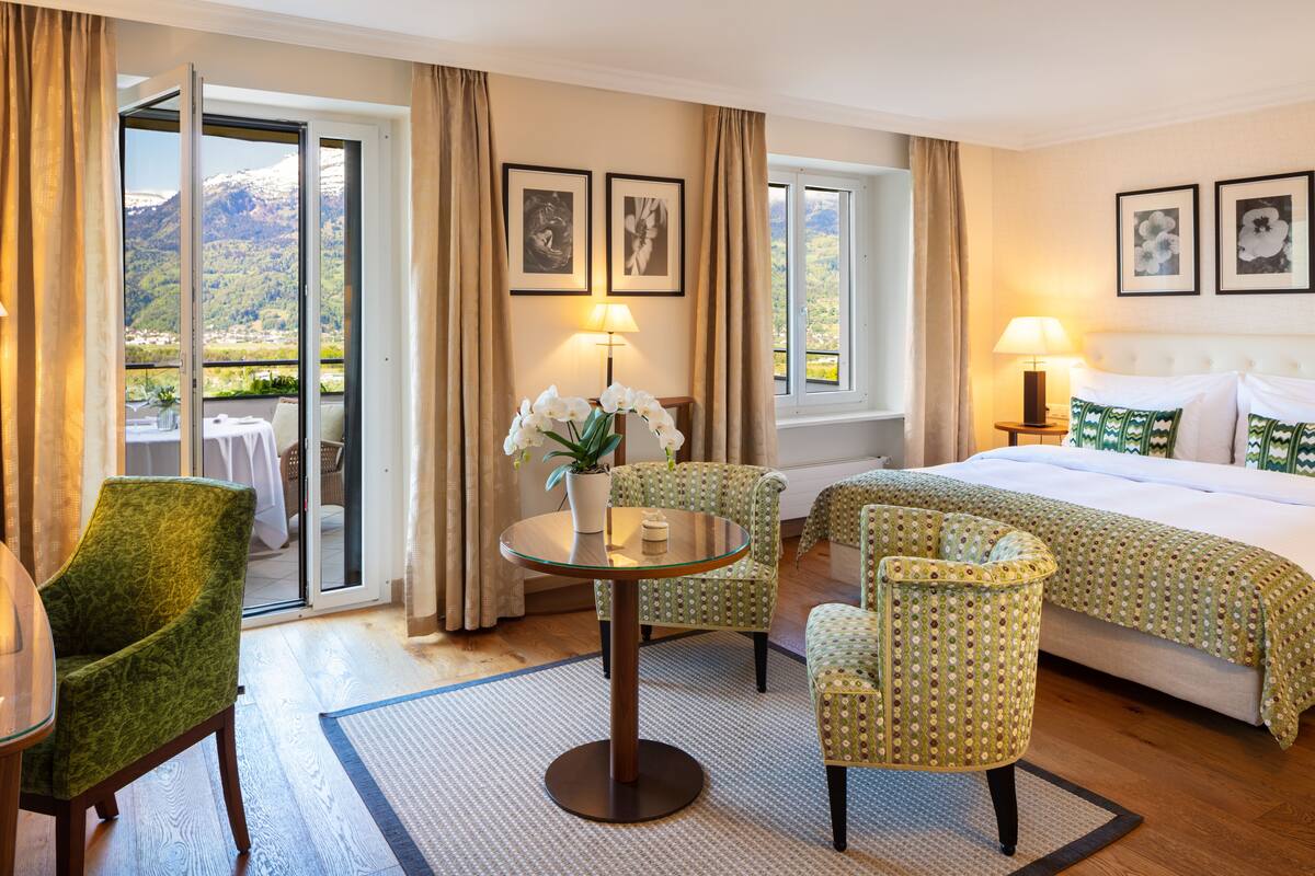 Een hotelkamer met een bed, twee stoelen, een tafel met een bloempot en uitzicht op bergen door de schuifdeuren.