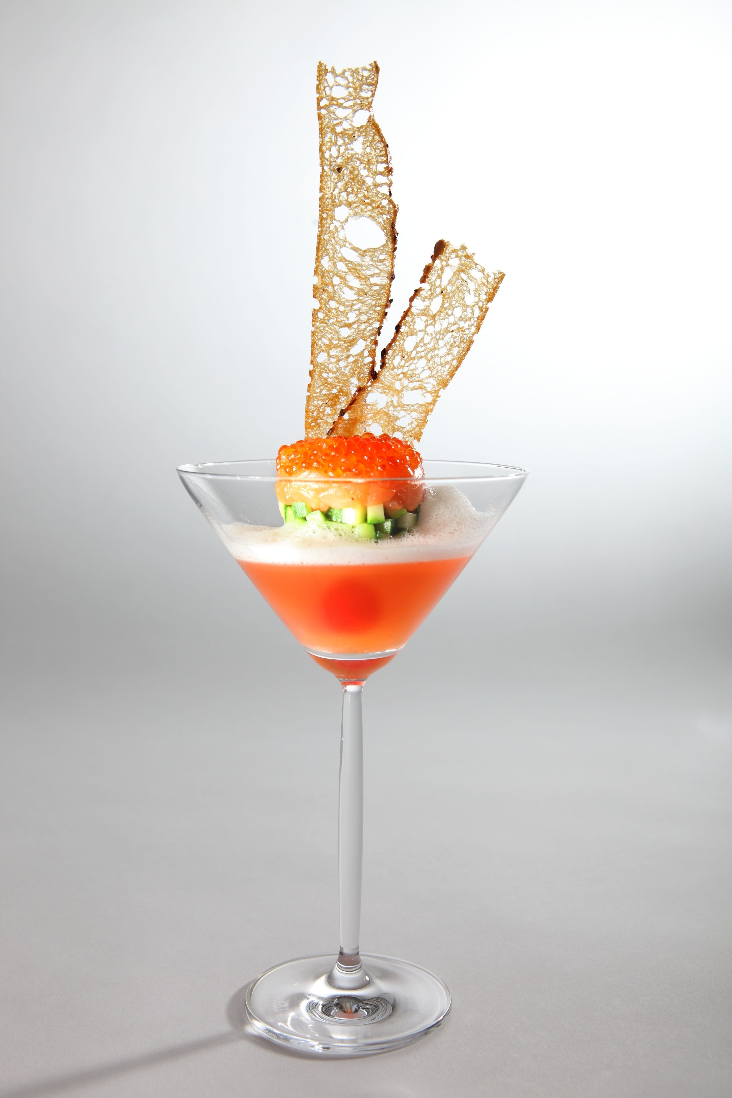A martini glass with a red cocktail, garnished with a caviar-topped ice sphere and crispy wafer sticks.