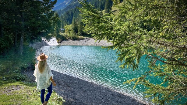 Une femme se promène sur un sentier de gravier qui longe un lac aux eaux turquoise et limpides, entouré d’une forêt verdoyante et dense, avec des montagnes en arrière-plan.