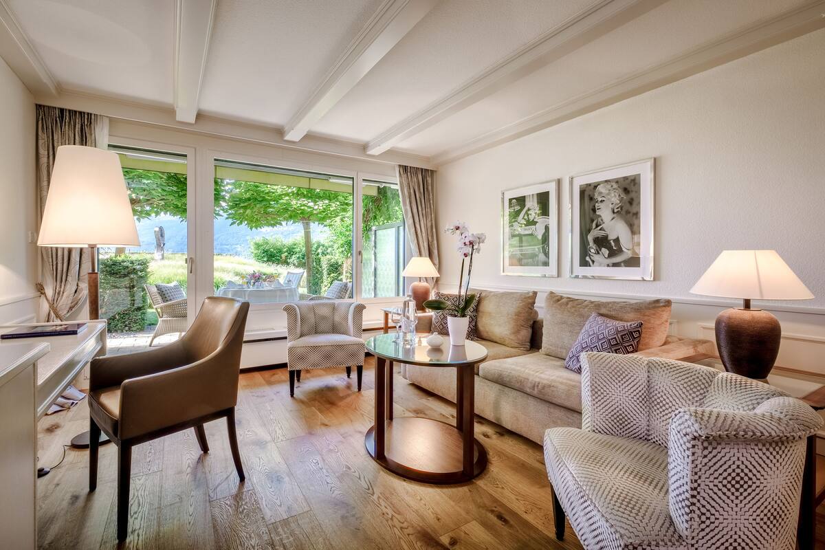 Een goed verlichte woonkamer met een houten vloer, een bank, twee fauteuils, een salontafel en een bureau met een lamp.