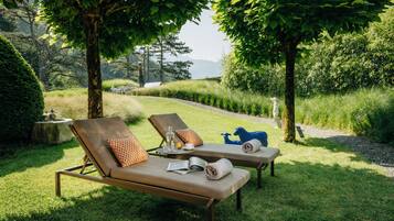 Deux fauteuils de relaxation, accompagnés de serviettes et d’un verre d’eau, sont disposés dans un espace extérieur verdoyant, agrémenté d’arbres et offrant une vue sur les collines.