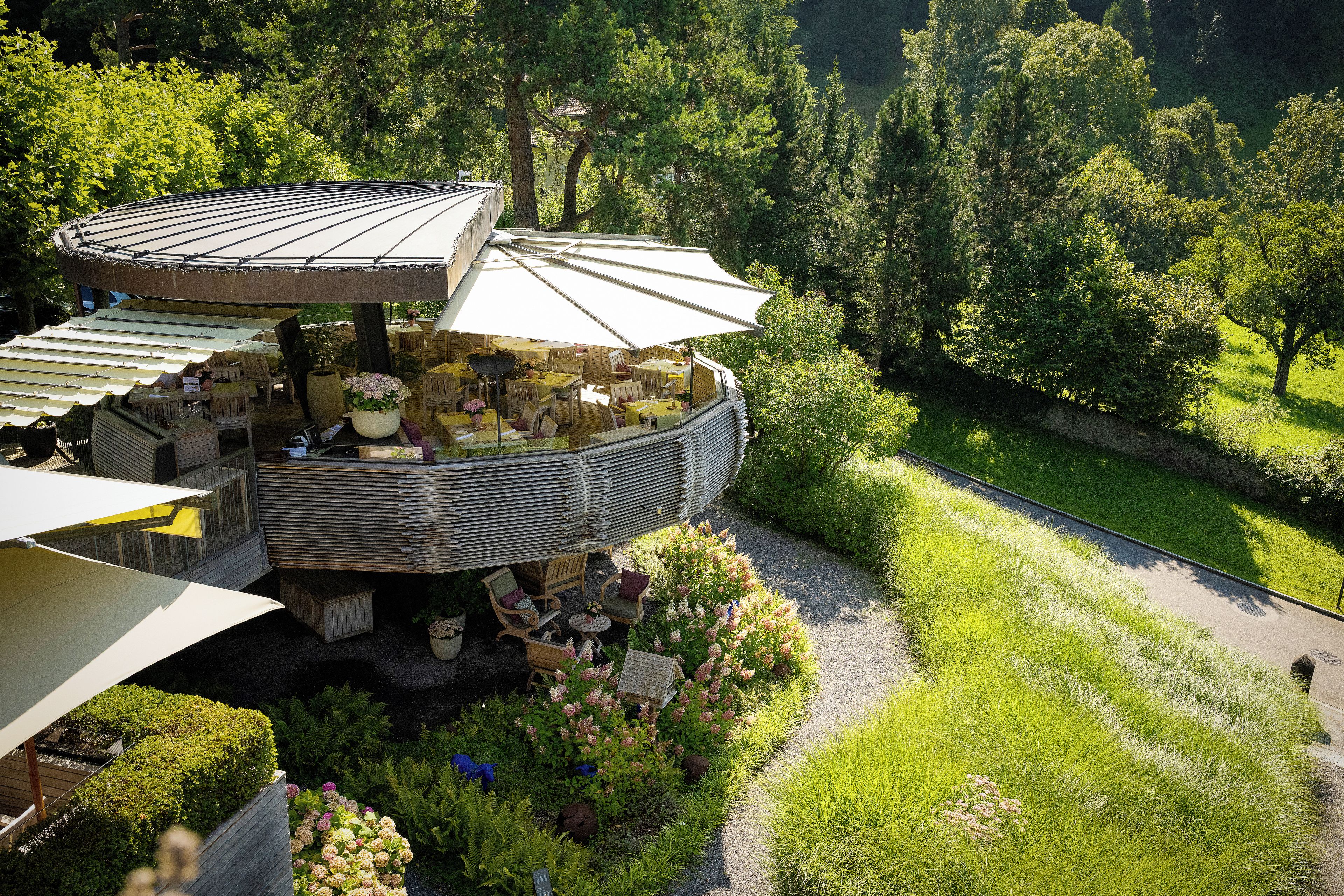 Un espace extérieur aménagé en plusieurs niveaux, avec des tables et des parasols, entouré d’une végétation luxuriante et d’arbres.