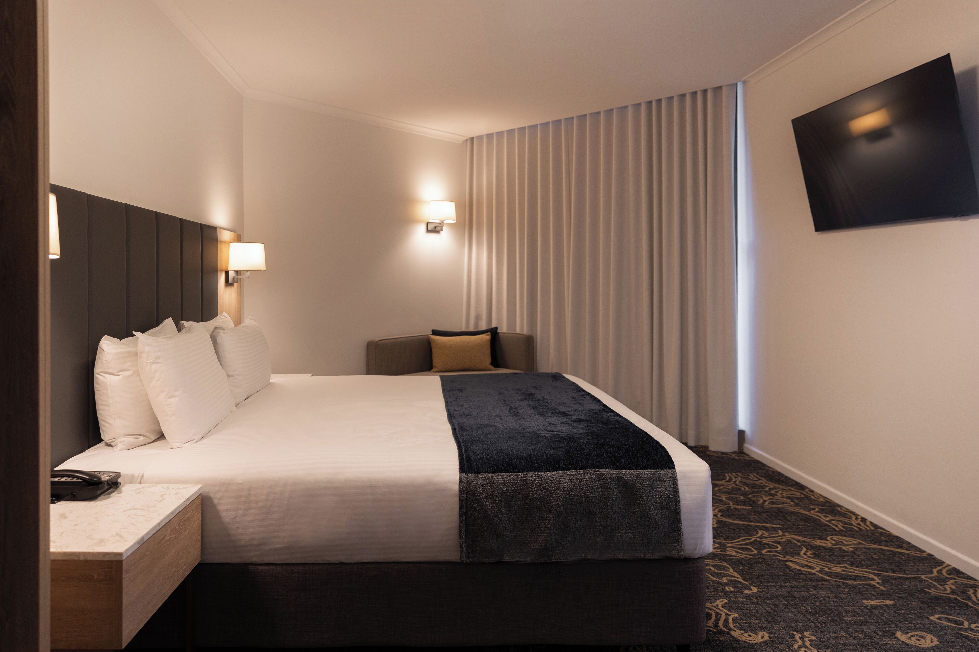 A hotel room with a bed, bedside table, lamp, and a flat-screen TV mounted on the wall.