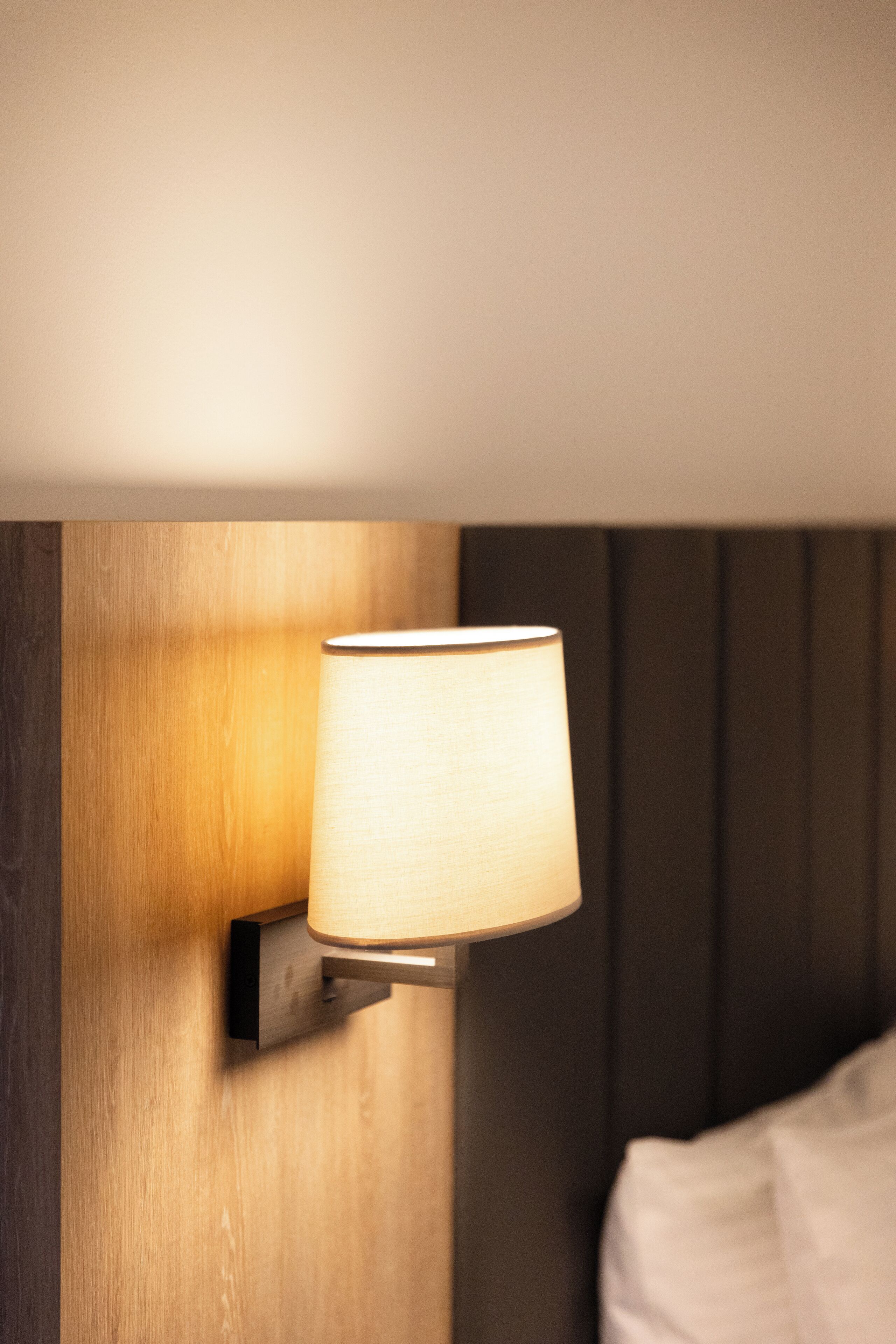 A wall-mounted lamp in a room with a wooden panel and a bed with white bedding.
