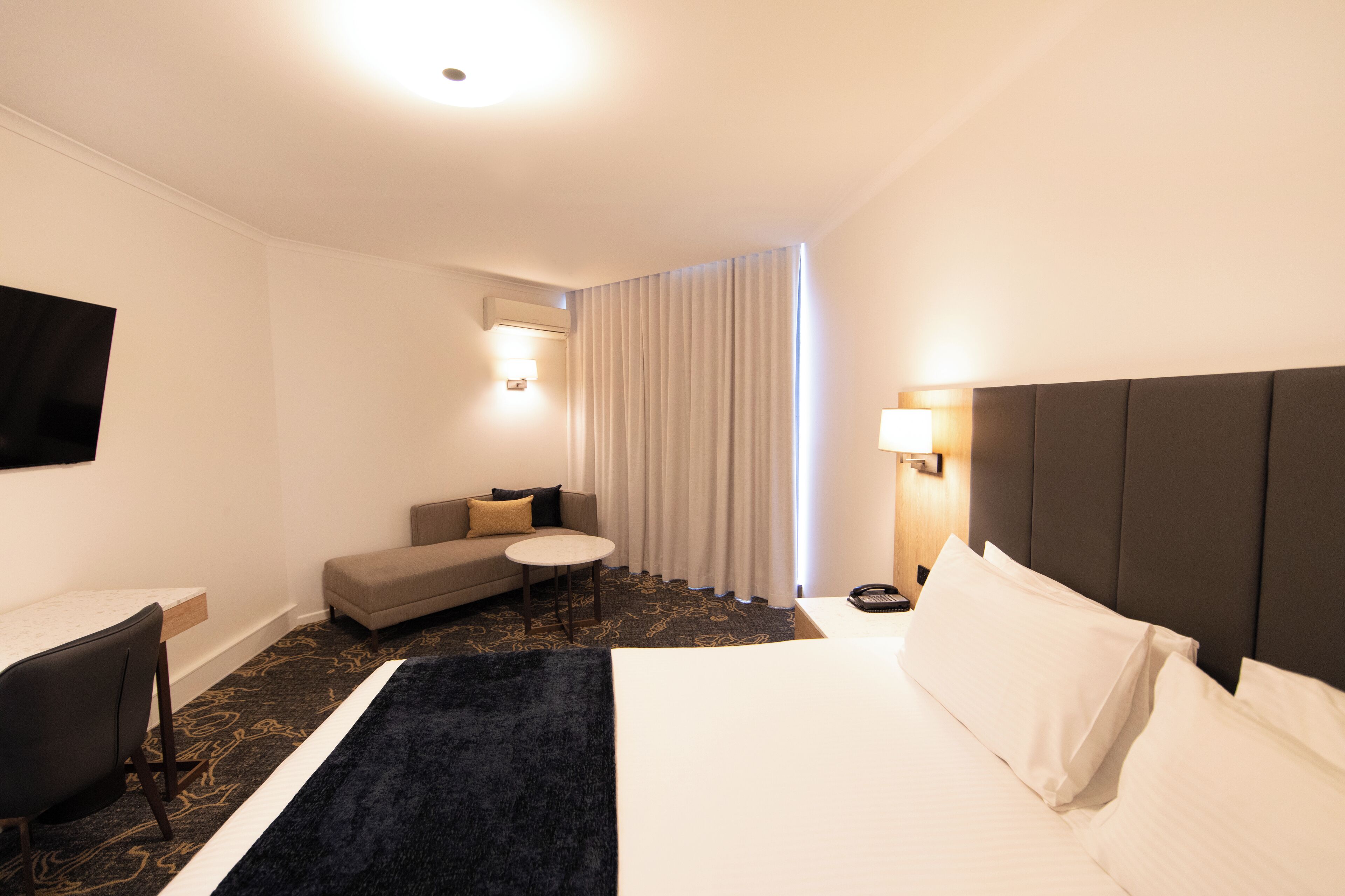Quarto de hotel com cama, sofá, mesa pequena, escrivaninha e televisão.