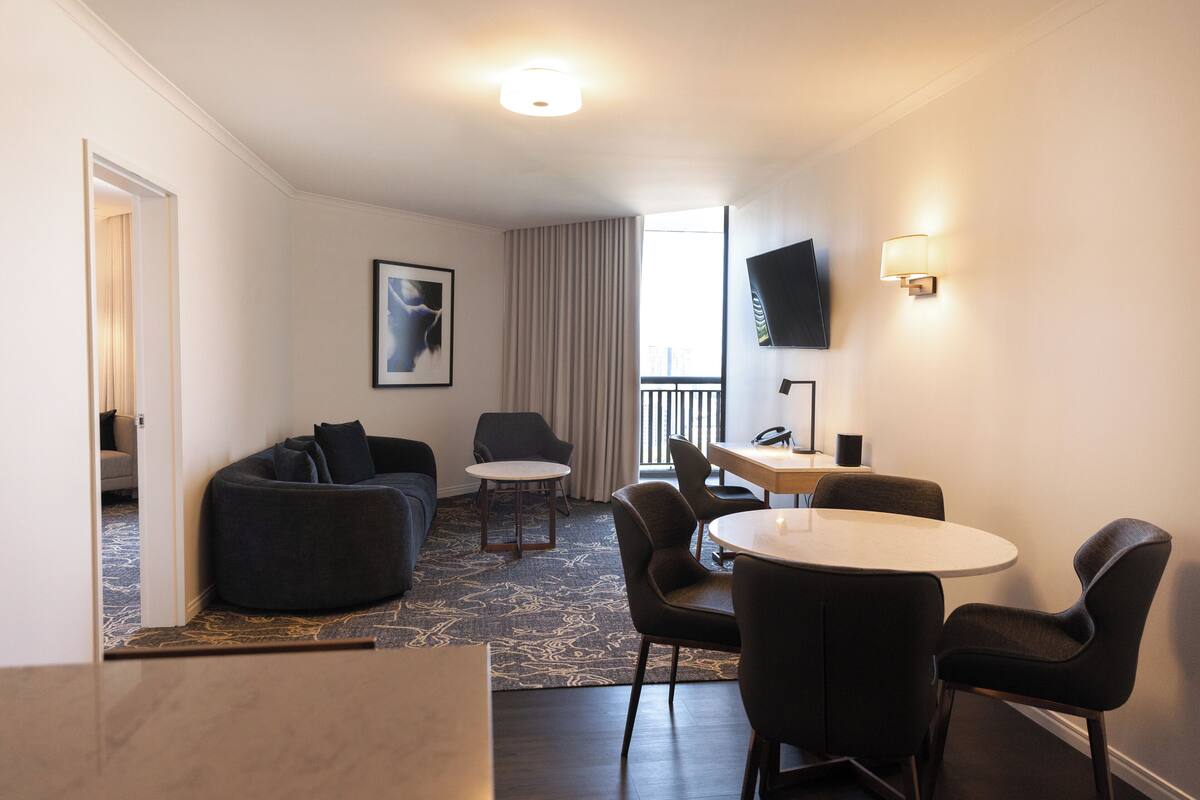 Een moderne hotelkamer met een zithoek, een eettafel met stoelen, een bank, een klein rond tafeltje, een flatscreen-tv aan de muur en een groot raam met gordijnen.