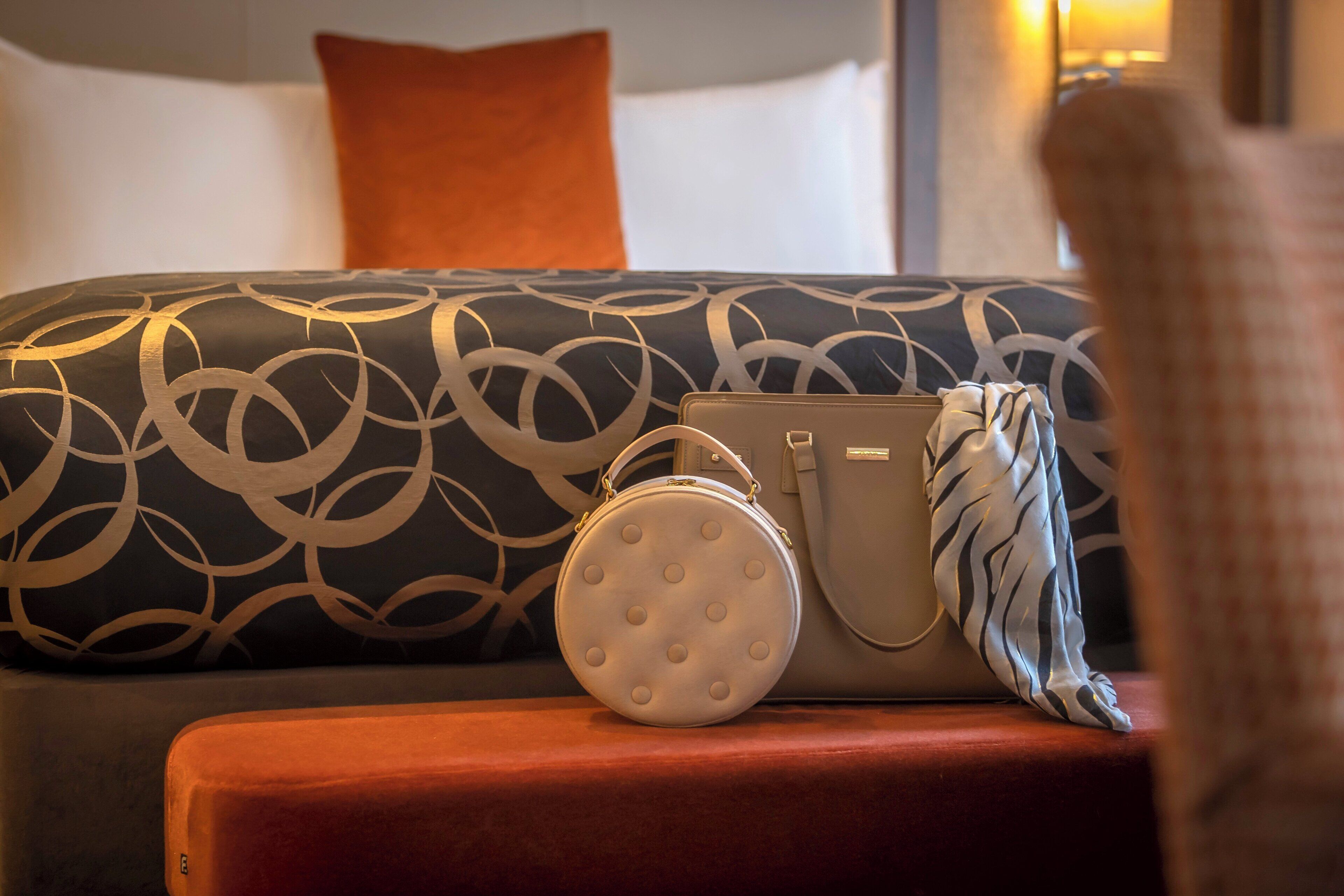 A bed with a patterned headboard, a round white pillow, and a beige handbag on a red ottoman.