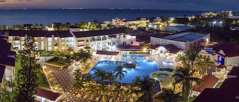 A resort with a large swimming pool, multiple buildings, and a view of the ocean.