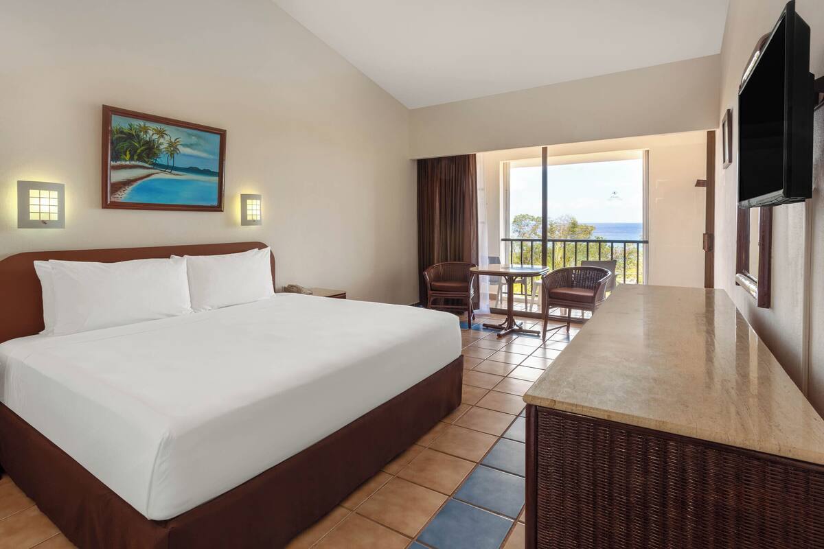 Une chambre d’hôtel avec un grand lit, un coin repas avec des chaises, une télévision et un tableau représentant une scène de plage.