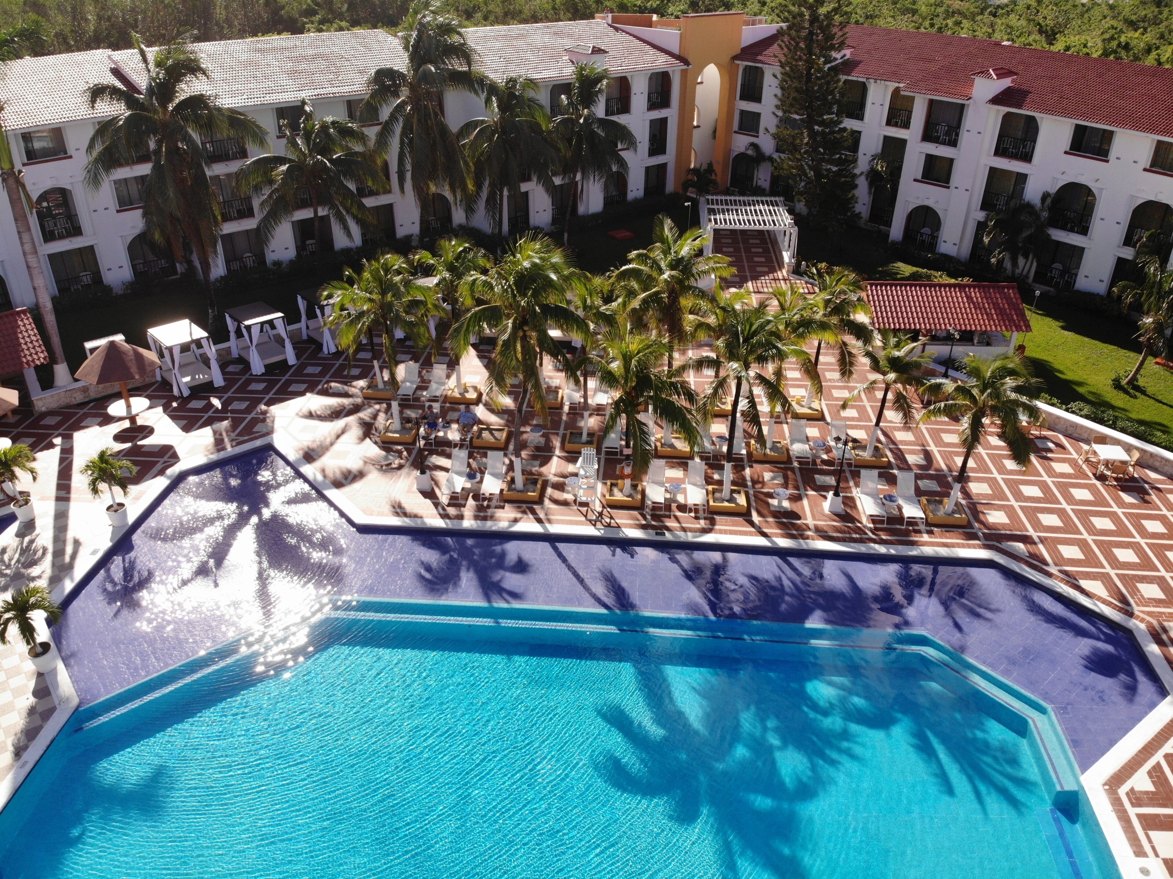 A resort pool area with multiple lounge chairs and palm trees, surrounded by a building with a tiled roof.