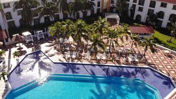 A resort pool area with multiple lounge chairs and palm trees, surrounded by a building with a tiled roof.