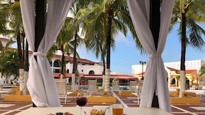 A dining area with a pool view, featuring a table with food and drinks, surrounded by palm trees and a building with red-tiled roofs. - Cozumel Hotel & Resort (Cozumel)