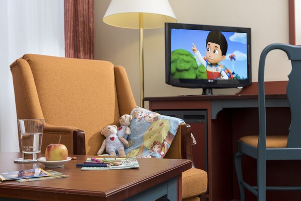 A hotel room with a large armchair, a television displaying an animated show, a small table with a glass of water, an apple, and a teddy bear.