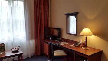 A hotel room with a desk, chair, television, lamp, and a window with curtains.
