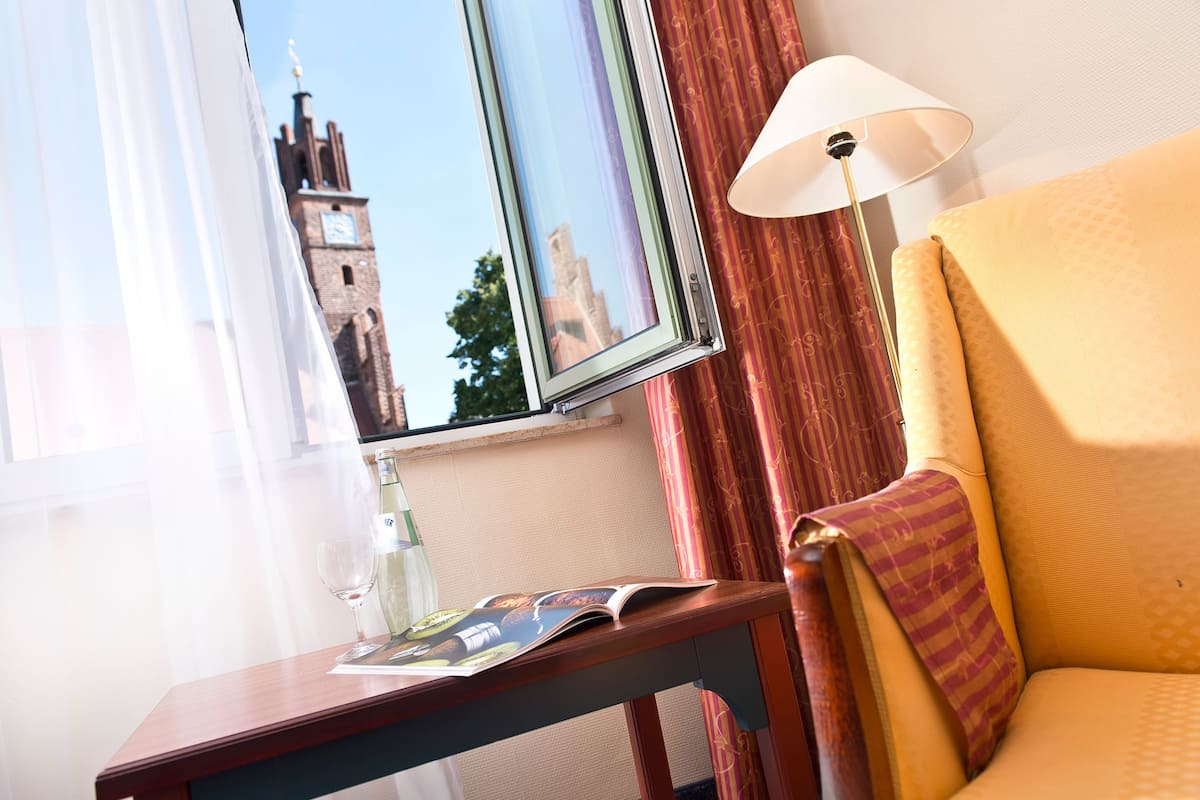 Une chambre d’hôtel avec une table en bois, un magazine, un verre d’eau, une lampe et une vue sur une tour, visible à travers la fenêtre.
