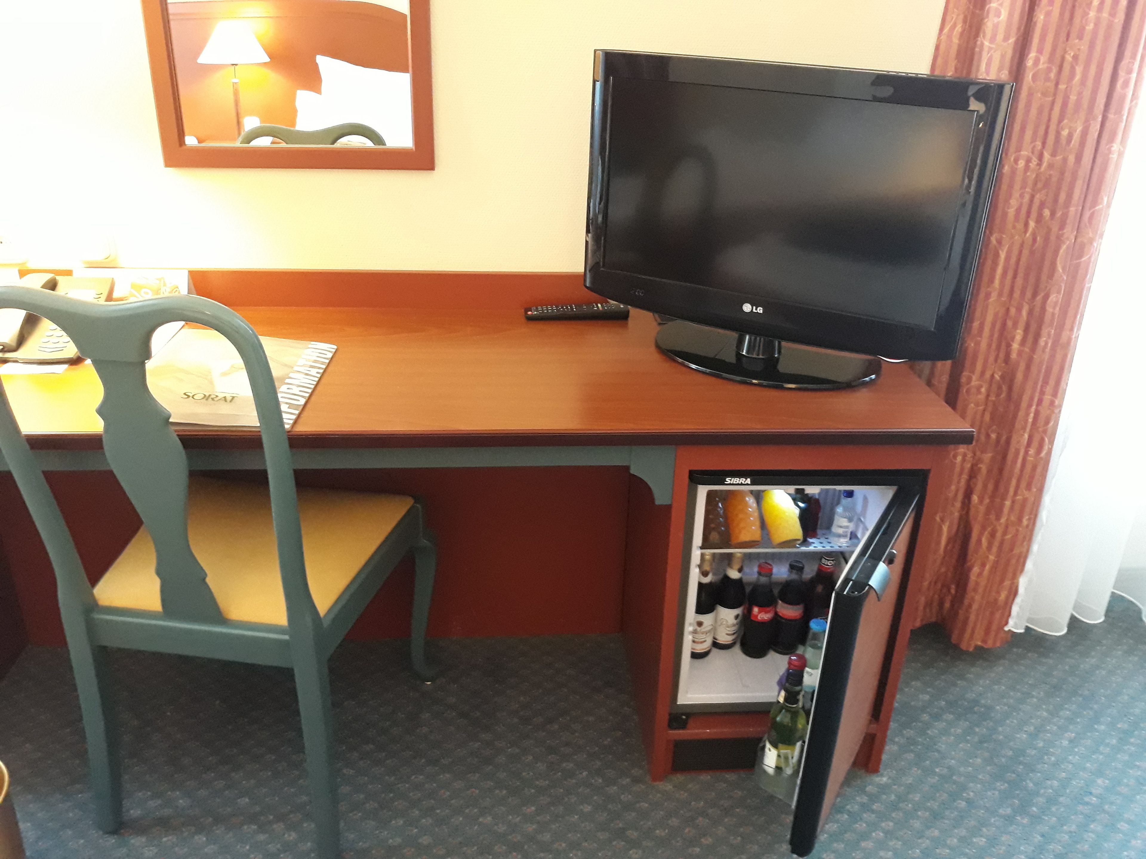 A hotel room with a wooden desk, a television, a chair, and a minibar.