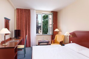 Ein Hotelzimmer mit einem Bett, zwei Stühlen, einem Schreibtisch mit Computer, einem kleinen Tisch mit Flasche und Gläsern sowie einem Fenster mit Vorhängen.