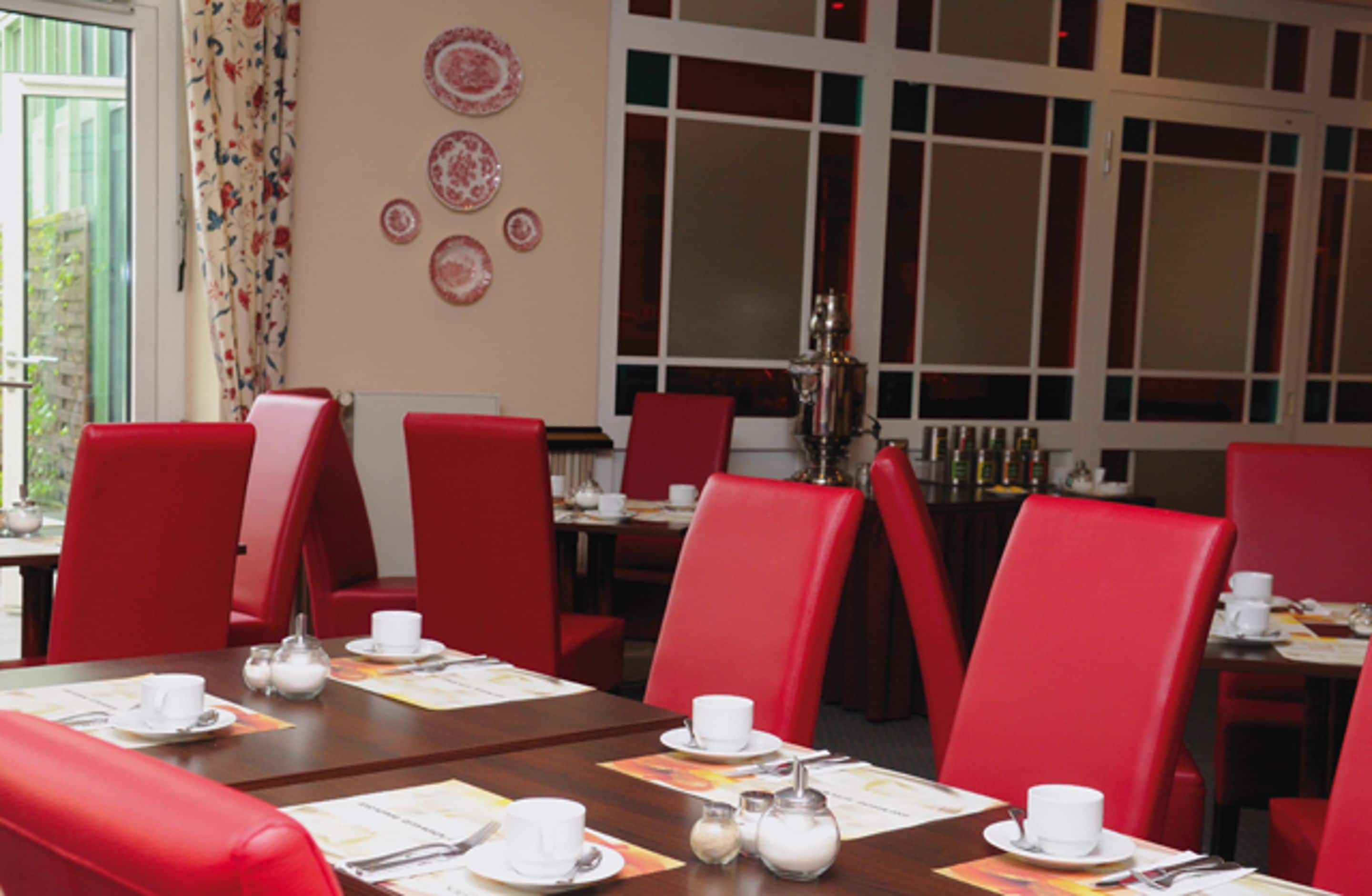 A dining area with red chairs and tables, a window with curtains, and a wall with decorative plates.