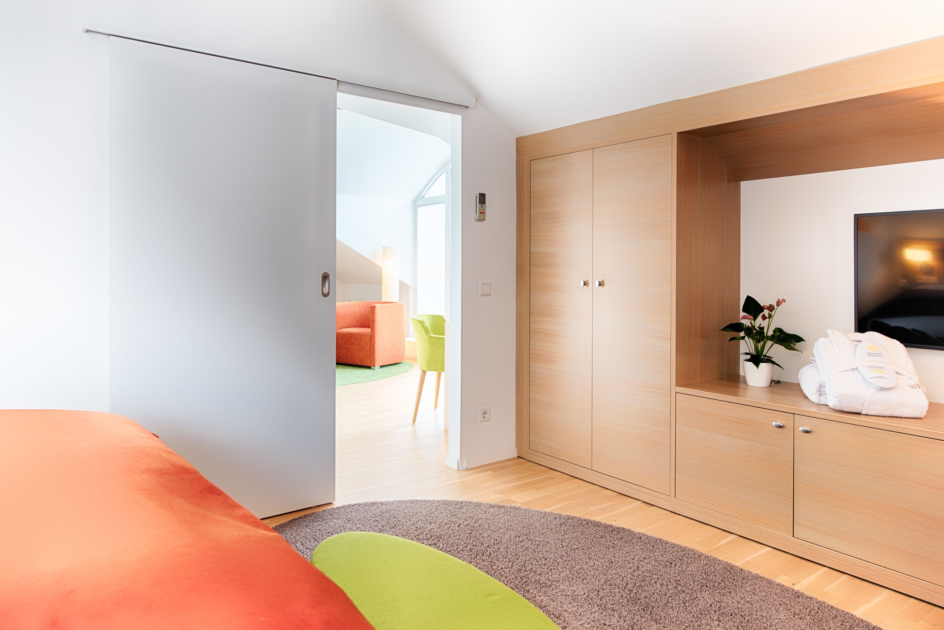 A modern hotel room with a wooden wardrobe, a flat-screen TV, and a view into a bright living area.