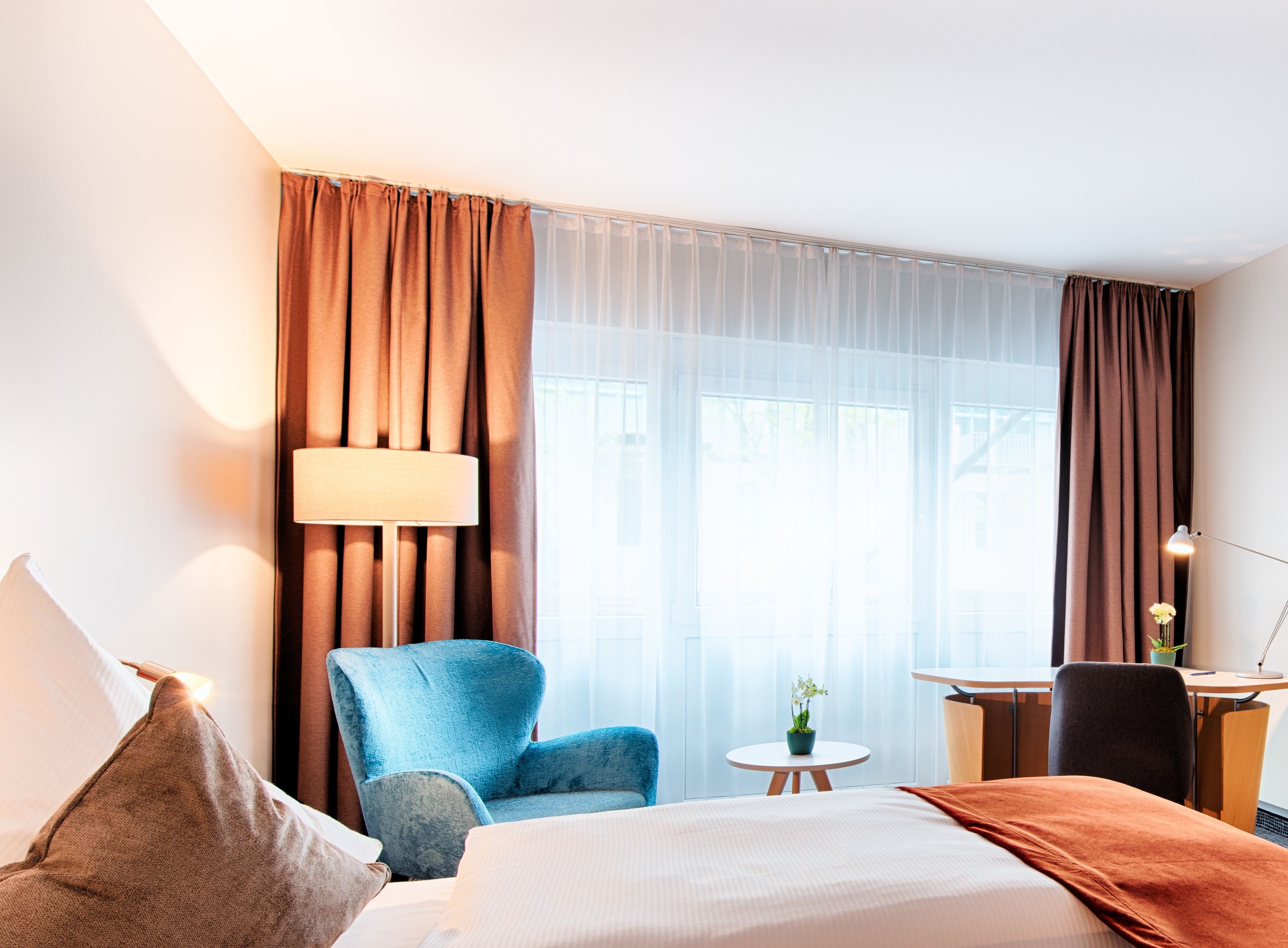 A hotel room with a bed, a blue armchair, a small round table with a plant, a desk with a chair, and a lamp.