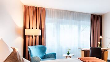 A hotel room with a bed, a blue armchair, a small round table with a plant, a desk with a chair, and a lamp.