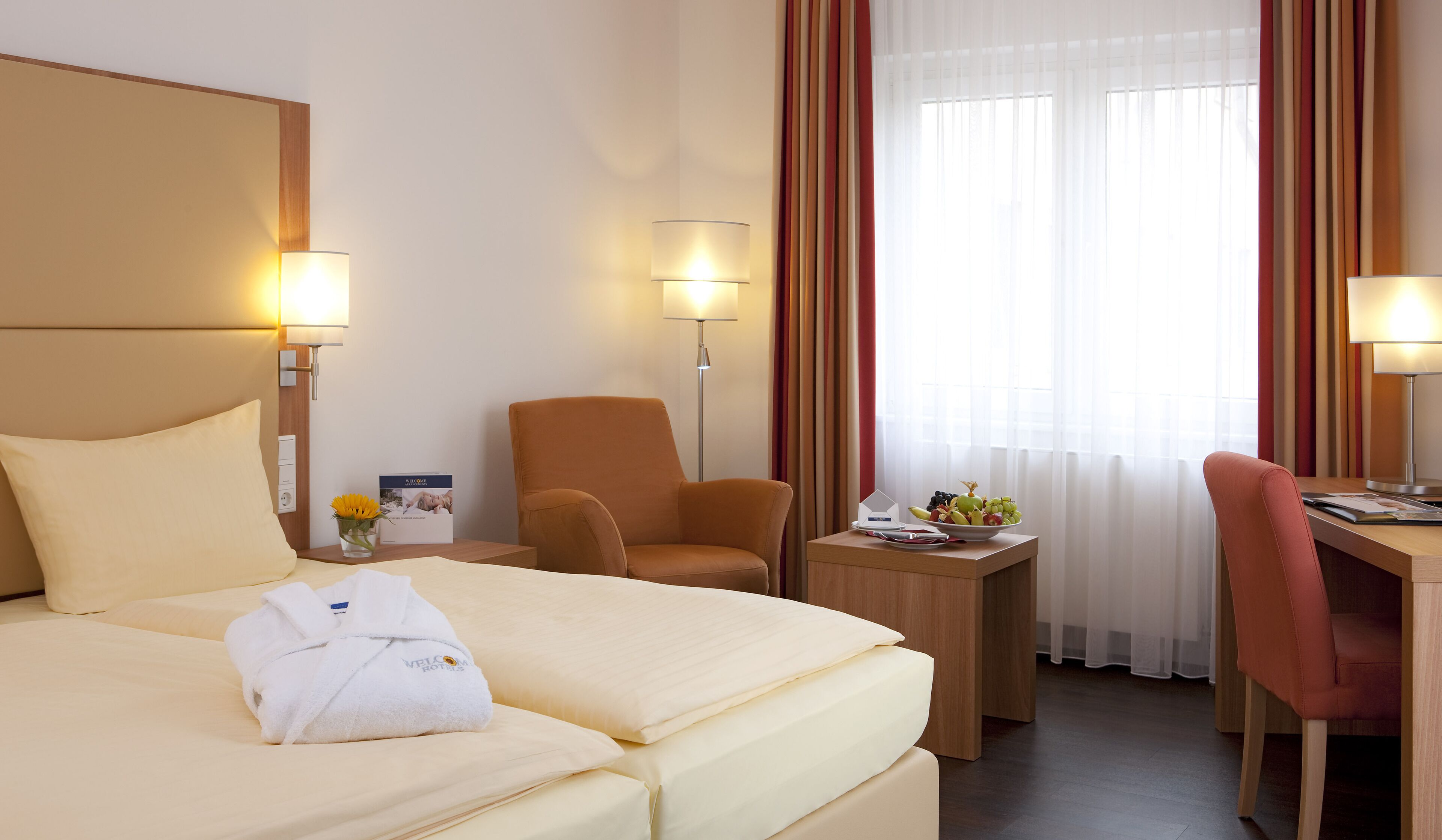 Une chambre d’hôtel comprenant un lit, deux chaises, un bureau, une petite table avec un panier de fruits et deux lampes.