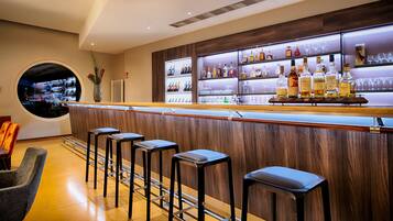 A bar area with a wooden counter, multiple stools, and shelves stocked with bottles and glasses.
