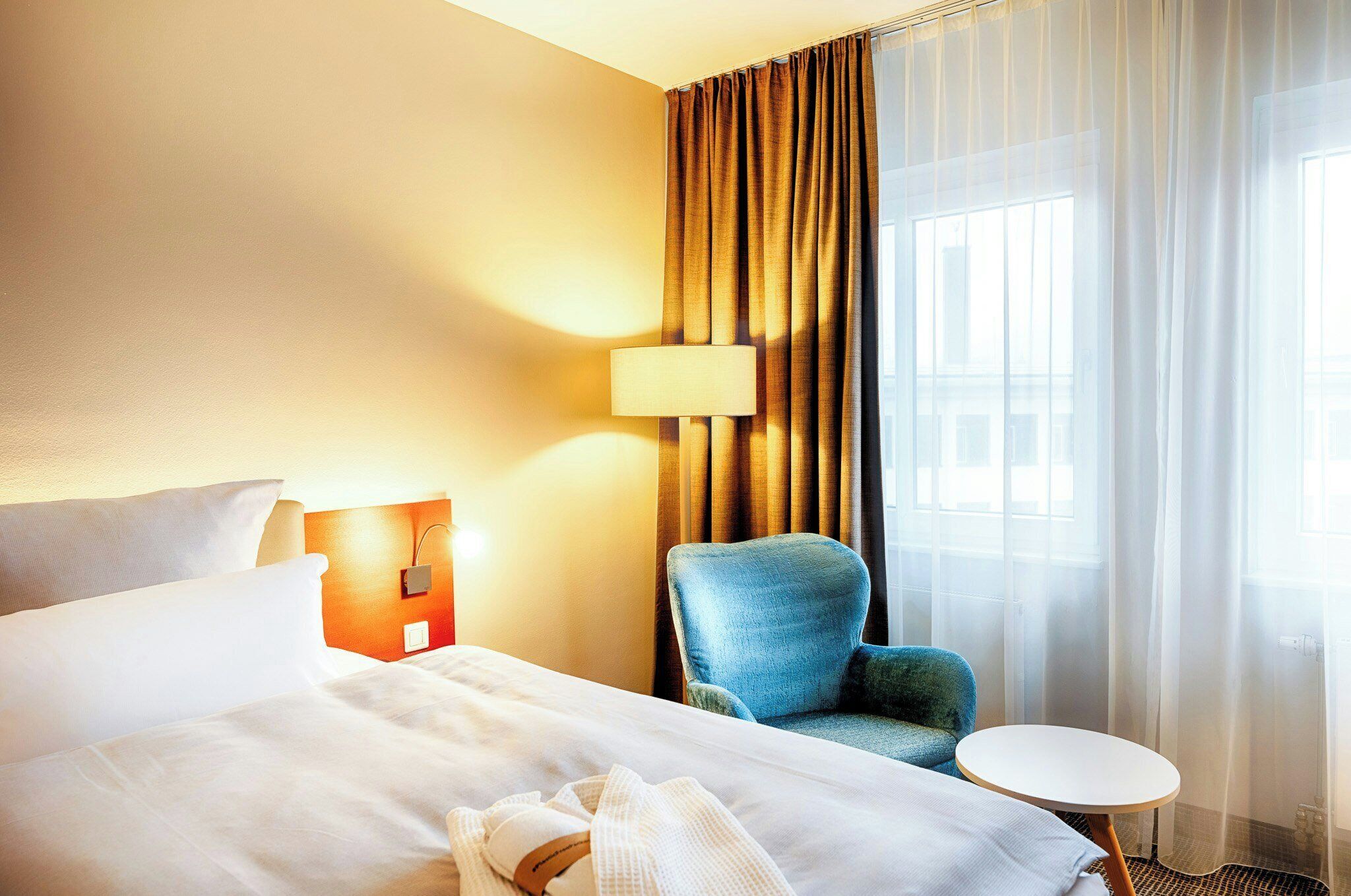 A hotel room with a bed, a chair, a small table, a lamp, and a window with curtains.