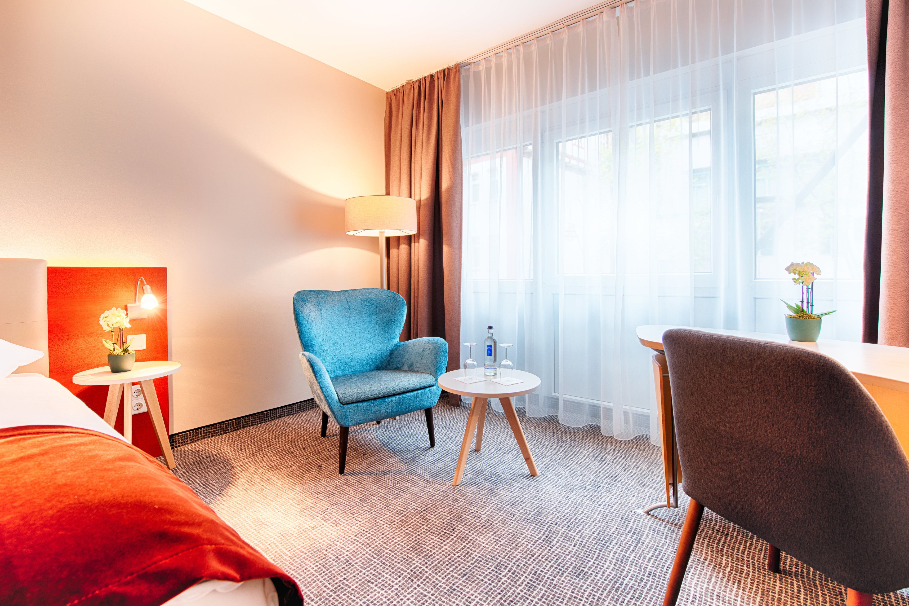 A hotel room with a bed, a blue armchair, a small round table with a lamp, and a view of the outdoors through sheer curtains.