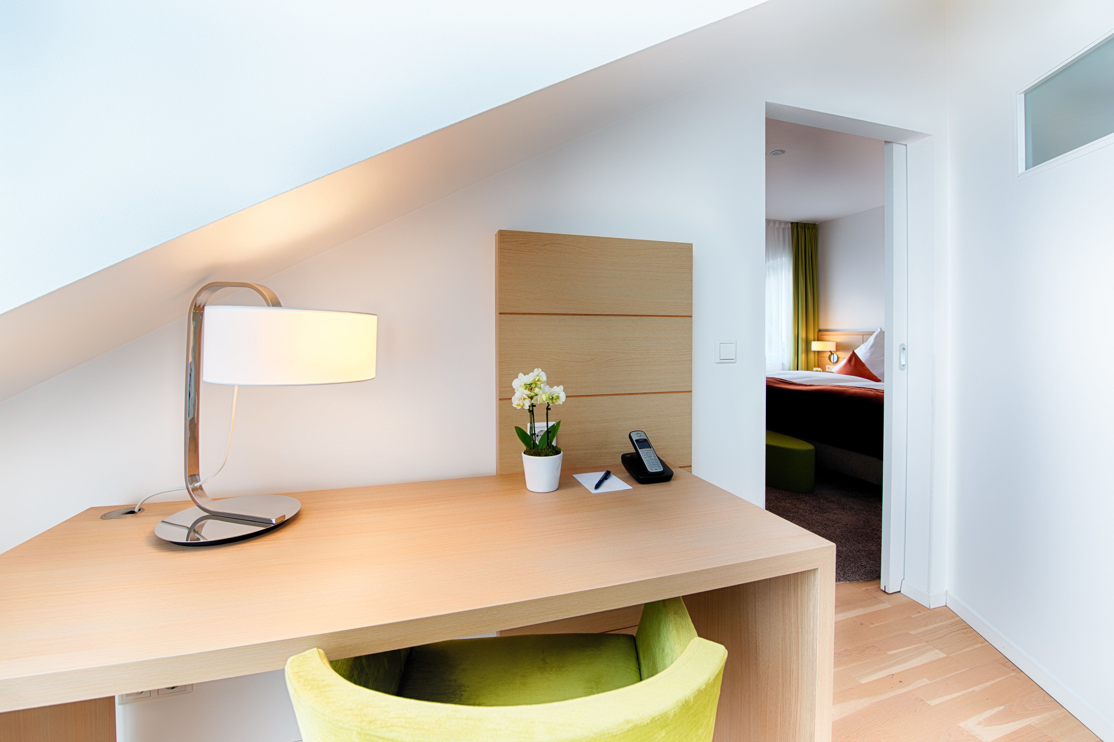 A modern hotel room with a desk, a lamp, a phone, and a small plant.