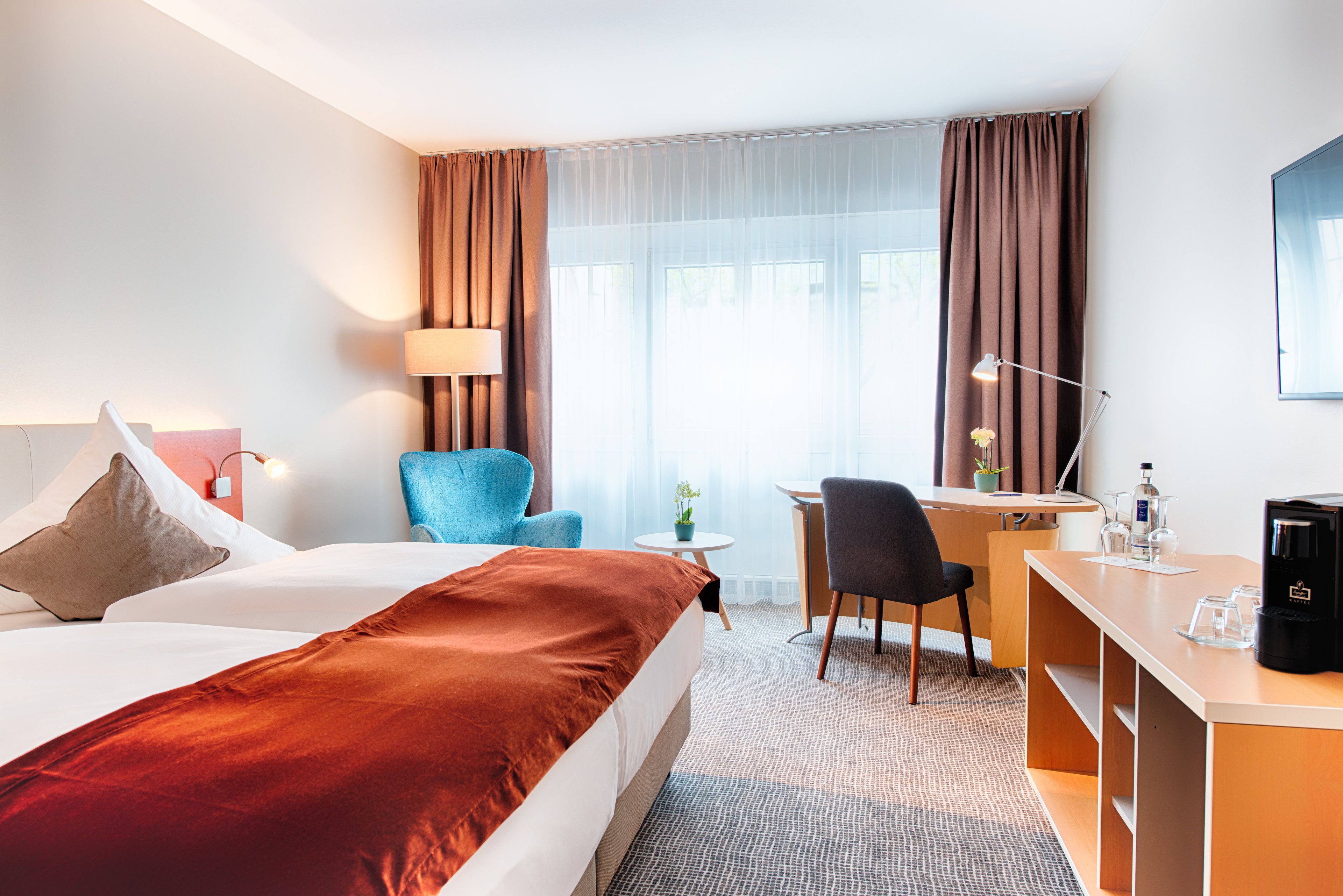 A hotel room with a bed, desk, chair, lamp, and a coffee machine.