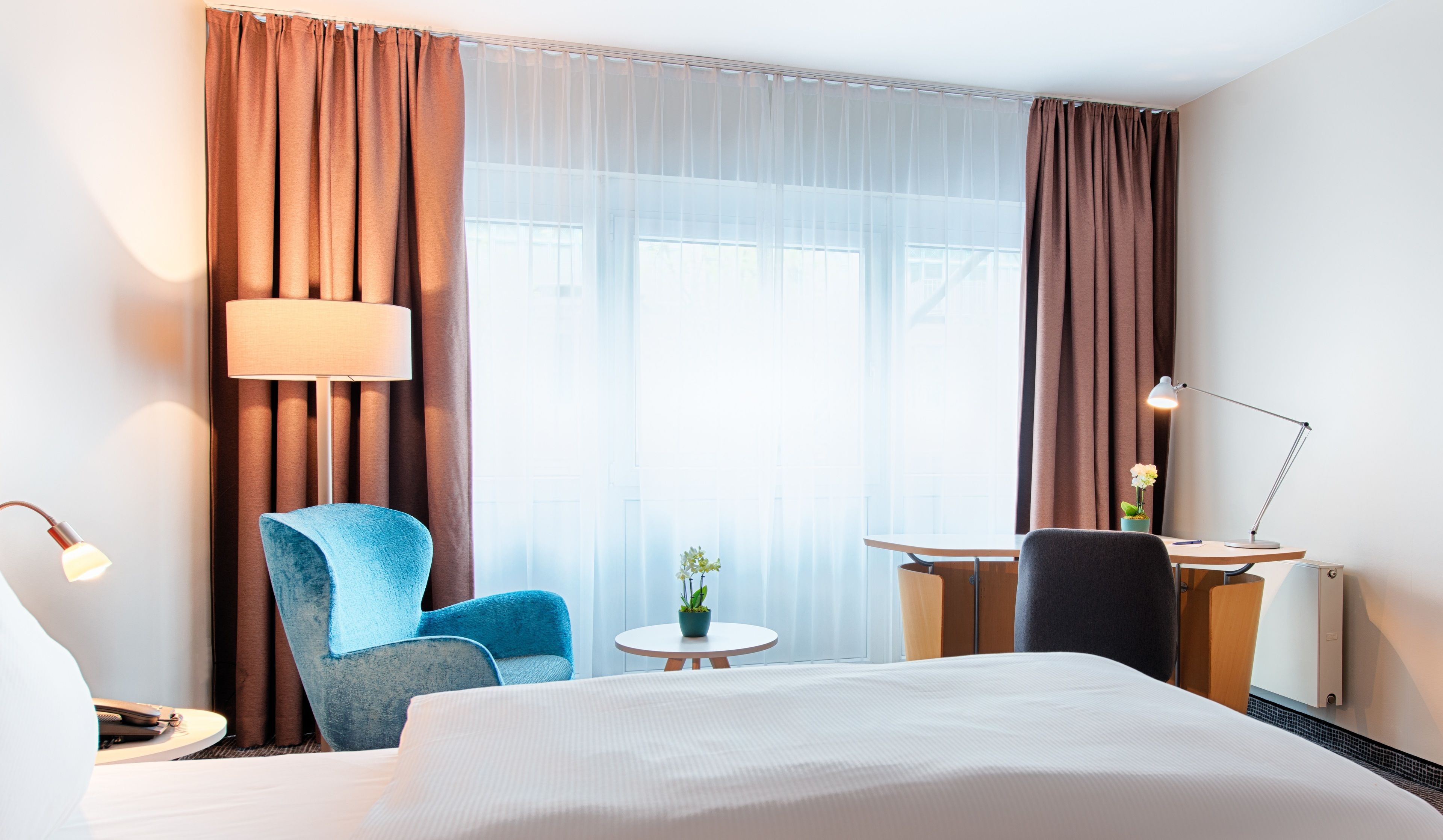 Una habitación de hotel con una cama, un escritorio con lámpara, una silla y una pequeña mesa con una planta.